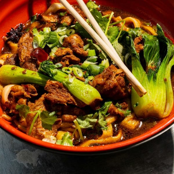 Beef noodle soup in a red bowl with bok choy, green onions, cilantro, and chopsticks.