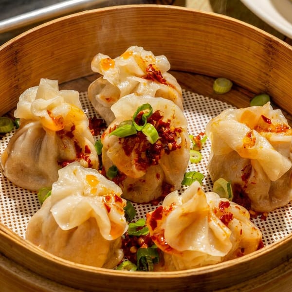 Six steamed dumplings topped with chili oil and green onions in a bamboo steamer.