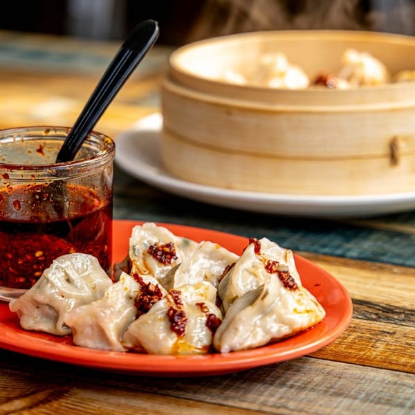 Steamed dumplings on a red plate with chili oil, and a bamboo steamer.