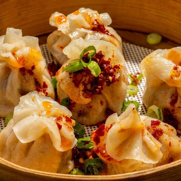 Steamed dumplings topped with chili oil and green onions in a bamboo steamer basket.